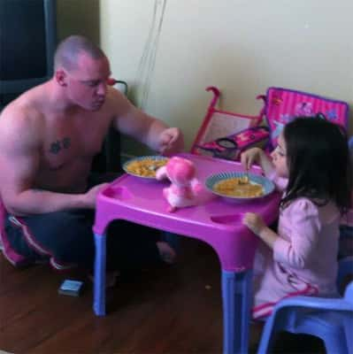 Pai do tamanho de um armário comendo na mesa de brinquedo da filha.