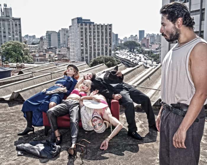 Em um telhado, ator Fernando Aveiro observa outros artistas da peça Ex-Gordo jogados em um sofá vermelho