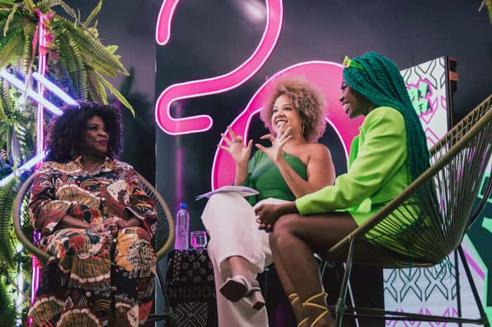 Na imagem, mulheres pretas se encontra em um palco, sorrindo e atentas a fala de outra mulher que se encontra com microfone em mãos enquanto fala.