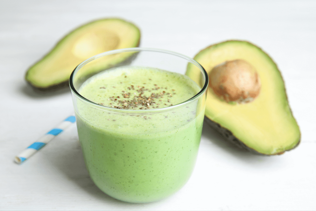 Copo transparente com vitamina verde coberta com sementes de chia, posicionado sobre uma superfície branca ao lado de metades de abacate e um canudo listrado de azul e branco.
