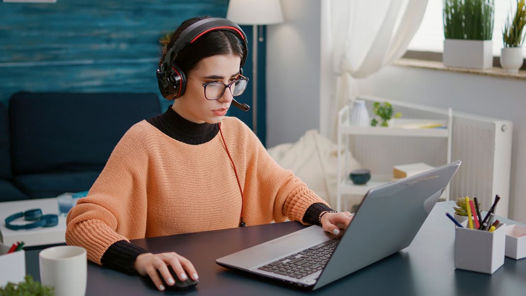 Young woman attending video call meeting on laptop with headphones, talking on remote teleconference for school class work. Female student using headset and computer on videoconference.