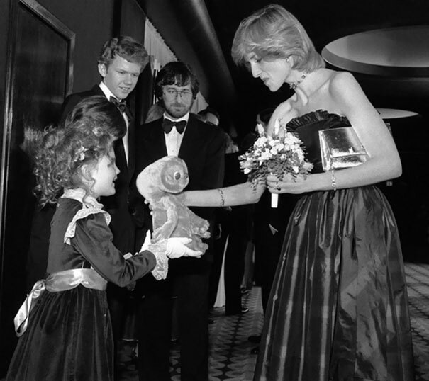 Drew Barrymore aos 7 anos de idade dando um ET de pelúcia à princesa Diana, enquanto Steven Spielberg e Robert Macnaughton observam a cena