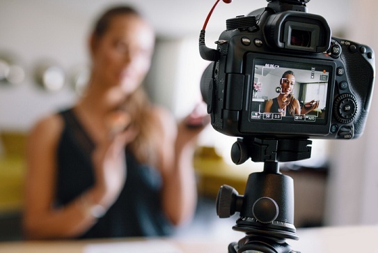 Woman making a video for her blog on cosmetics using a tripod mounted digital camera. Young female blogger on camera screen holding cosmetics.