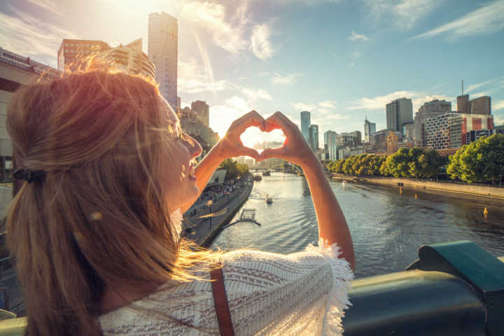 Melbourne makes a heart shape finger frame. She is standing on a bridge overlooking the Melbourne CBD along the Yarra River on a beautiful day.