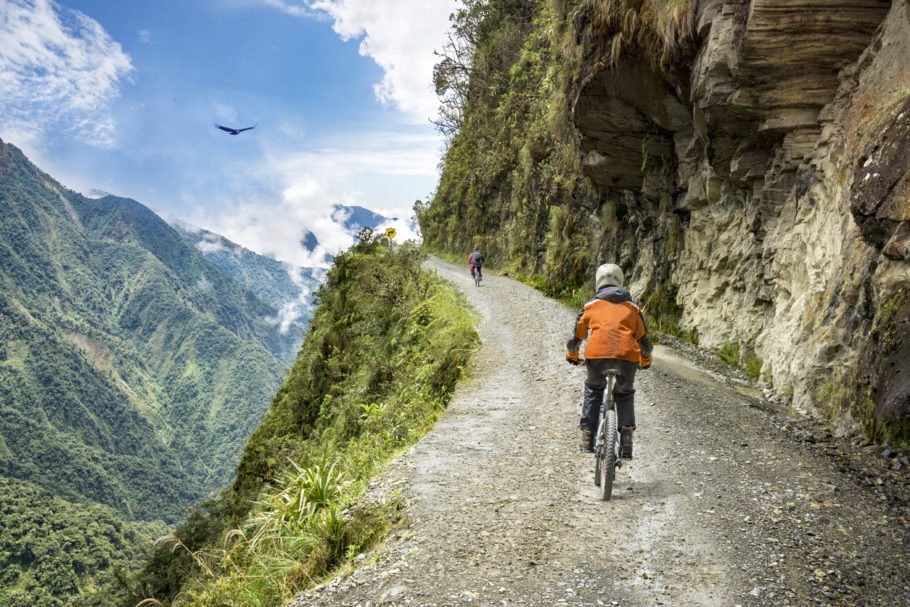 Bike adventure travel photo. Bike tourists ride on the “road of death” downhill track in Bolivia. In the background sky circles a condor over the scene.