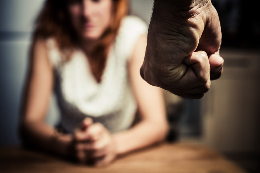 foto com uma mulher desfocada sentada à mesa com as mãos juntas e um punho masculino fechado focado e demonstrando raiva à mulher