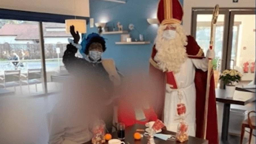 Idosos desfrutando de vinho quente e laranjas enquanto o Papai Noel e Zwarte Piet (“Peter Negro”) visitam asilo na Bélgica