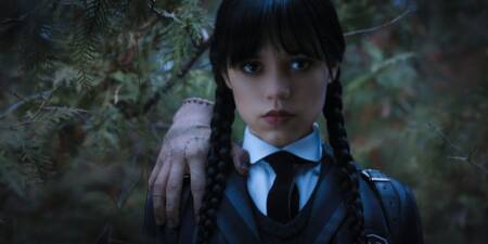 Cena mostra a atriz Jenna Ortega interpretando Wandinha Addams. Ela está séria, usa trancinhas no cabelo e está com o Mãozinha no ombro