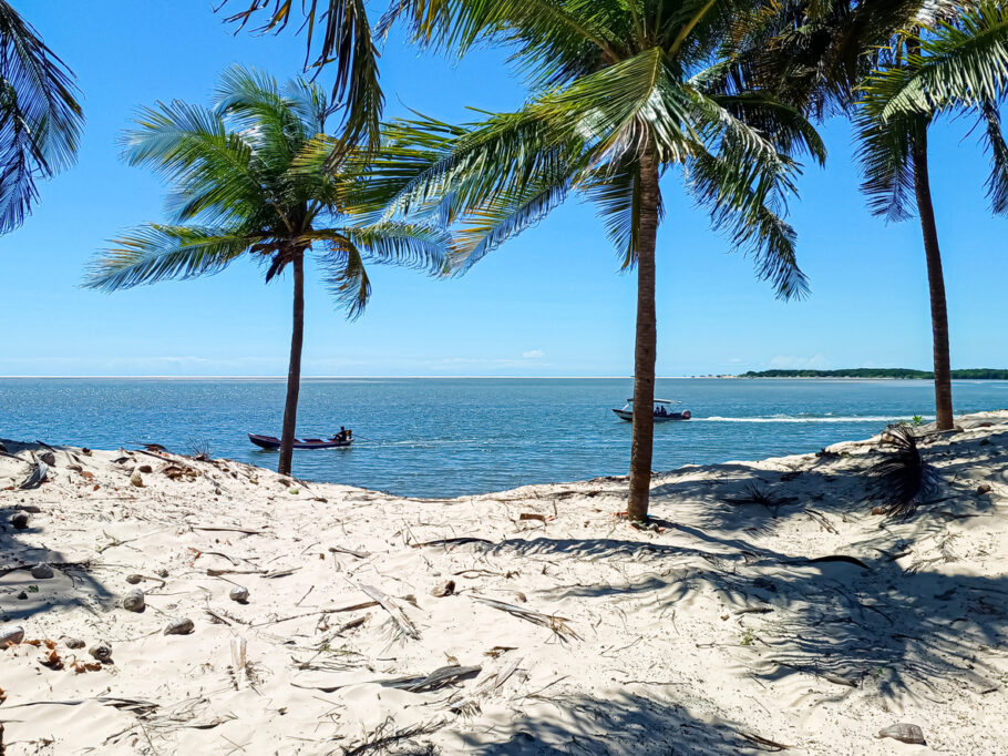 Praia de Atins nos Lençóis Maranhenses Brasil, vista com coqueiros e barcos com pessoas no mar azul sob o sol forte.