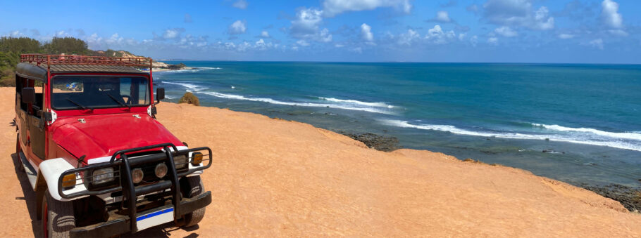 Foto panorâmica da praia de Pipa no estado do Rio Grande do Norte, foto de um carro vermelho em terreno arenoso, vista do mar e da orla