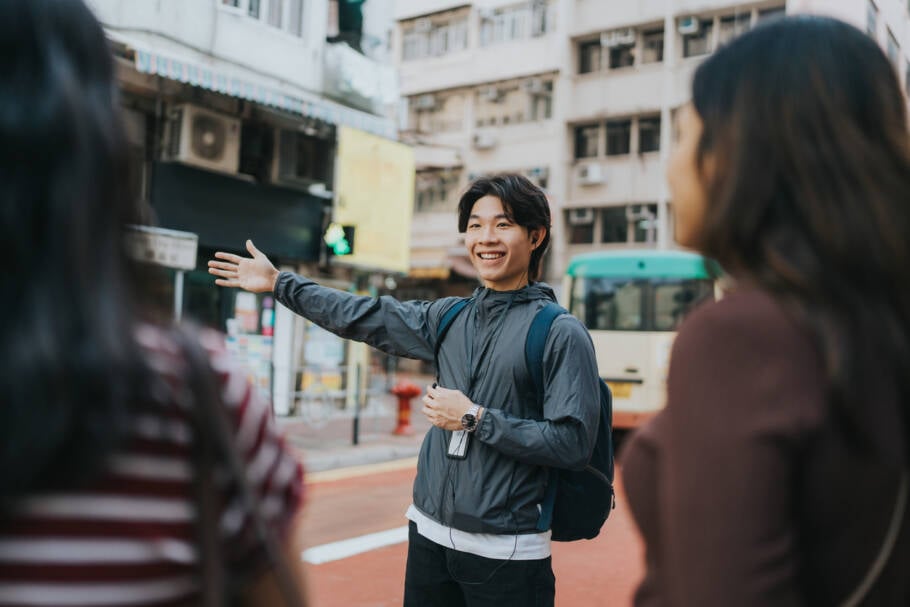 A medium shot of a Chinese tour guide pointing directions to the tourists while using an earphone and carrying a bag pack. They are in Ap Lei Chau Street with restaurants and buildings along the street.