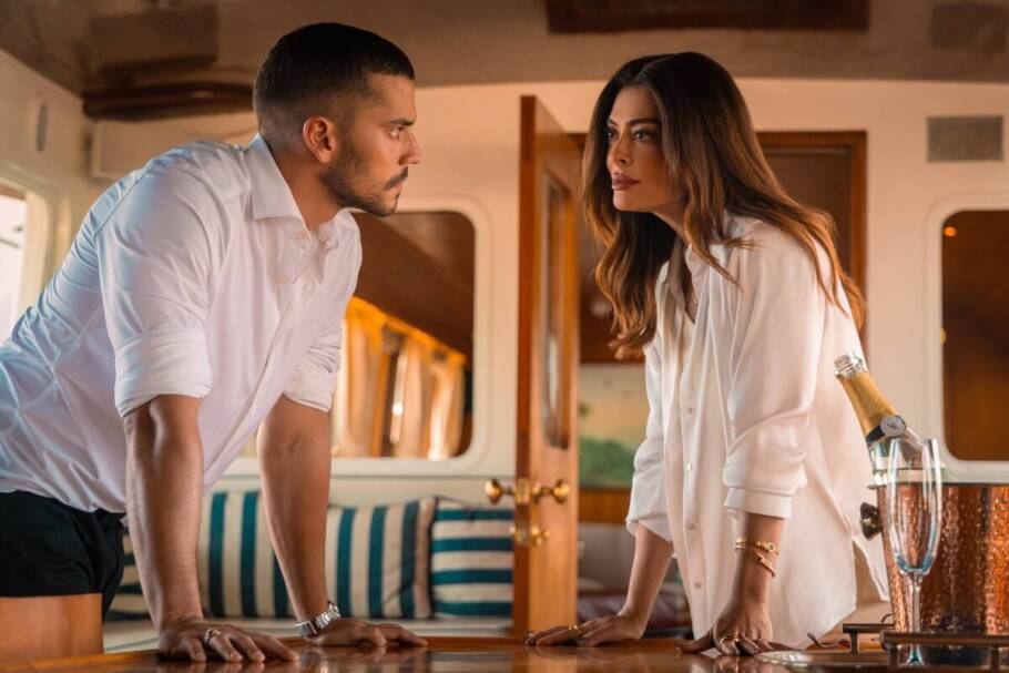 André Lamoglia e Juliana Paes inclinas e apoiados sobre uma mesa, conversando, em cena da série "Os Donos do Jogo"