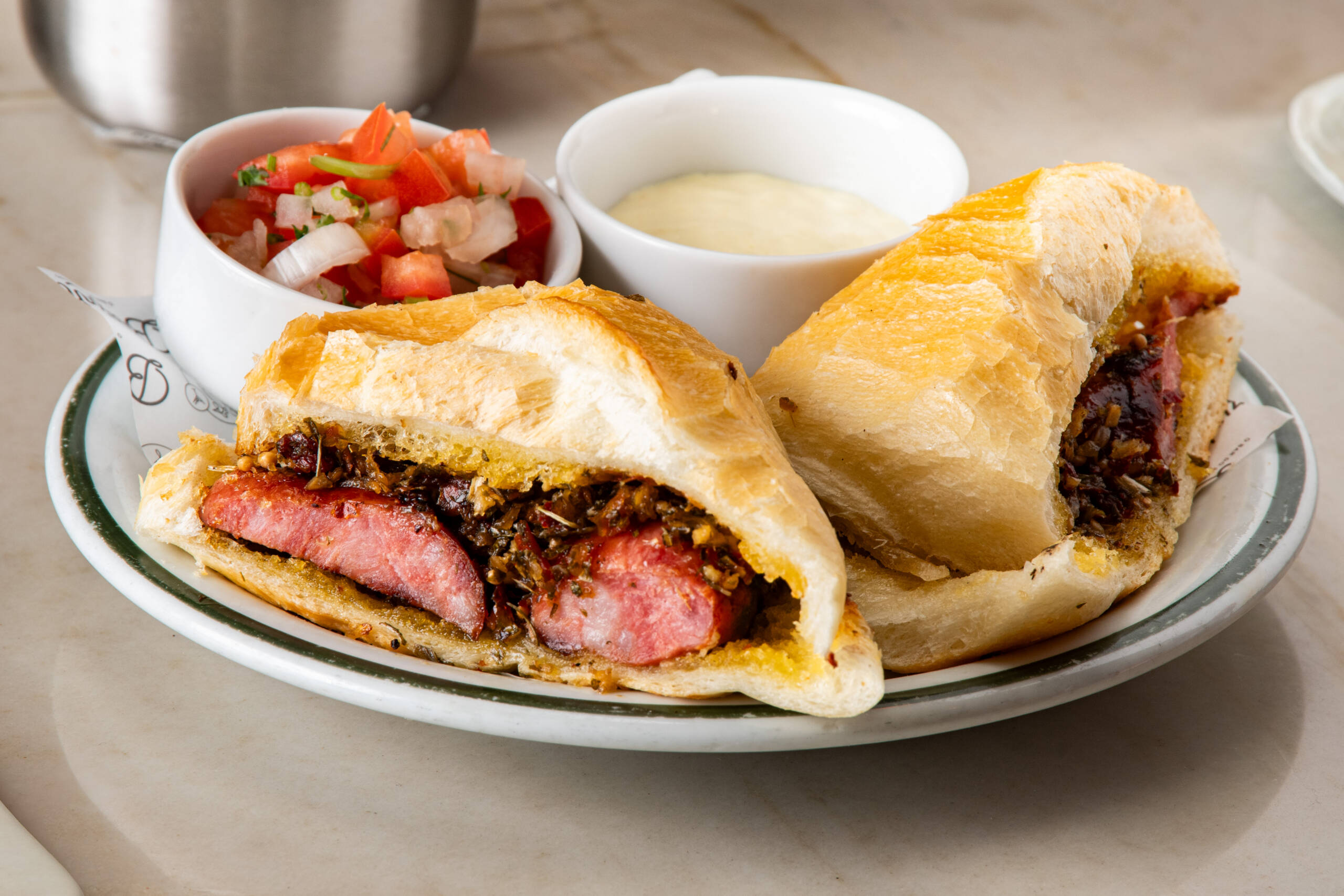 No Brazinha o choripán segue fiel à tradição: a linguiça vai direto para a brasa, chega ao pão francês ainda quente e recebe chimichurri na medida certa