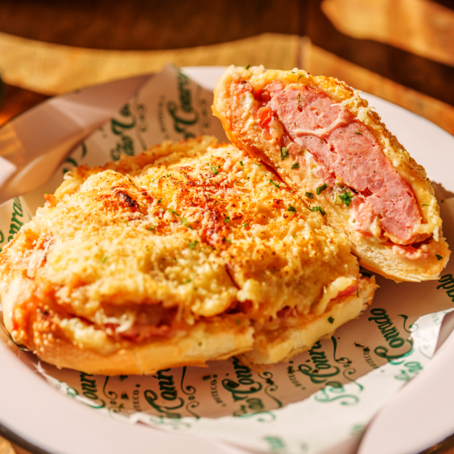 No São Conrado Bar o clássico ganha uma leitura mais brasileira servido no pão francês. Além da linguiça suculenta, entram tomate ralado, maionese e uma camada generosa de queijo gratinado, que derrete e domina o visual do sanduíche