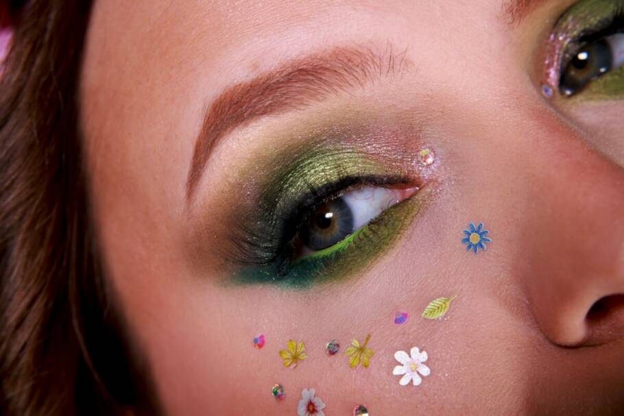 Fotografia do rosto de uma mulher, com foco na pálpebra, que apresenta um degradê de sombras verdes cintilantes e tons terrosos, com um delineado esfumado e cílios longos. Abaixo do olho e na maçã do rosto, há diversas aplicações decorativas em miniatura, incluindo pequenas flores brancas e azuis, folhas verdes e cristais de strass brilhantes, criando um efeito de "sardas artísticas". A pele é clara e a sobrancelha é definida em um tom castanho quente.