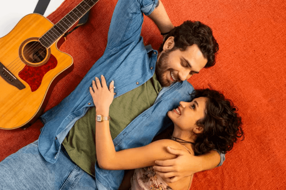 Homem com cabelo curto, barba, usando camiseta verde e jaqueta e calça jeans abraçado com mulher com cabelo curto usando regata de alcinha com violão ao lado do homem em fundo laranja