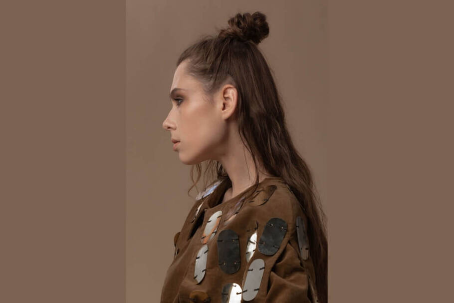 Fotografia de perfil de uma mulher com cabelos castanhos longos e ondulados, usando um penteado "half bun" (meio coque) alto e texturizado no topo da cabeça, com a metade inferior dos fios solta sobre as costas. Ela veste uma jaqueta marrom de camurça decorada com grandes aplicações ovais metálicas e reflexivas. A composição possui tons terrosos, com um fundo liso em bege escuro e iluminação suave que destaca o contorno do seu rosto e a textura do cabelo.