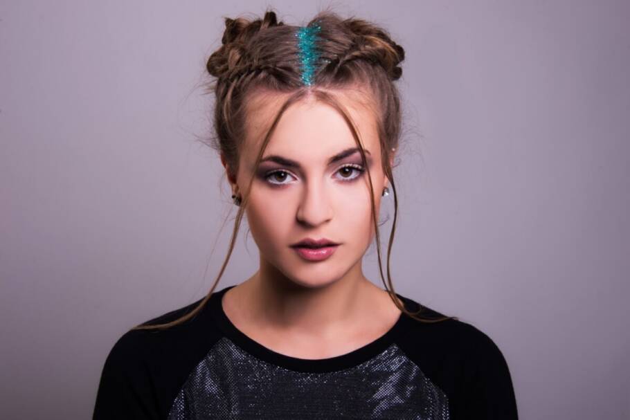 Retrato frontal de uma mulher jovem comO cabelo está dividido ao meio e preso em dois coques trançados nas laterais do topo da cabeça (estilo "space buns"), com a risca central da raiz preenchida por glitter azul-turquesa vibrante. Ela possui mechas finas de cabelo soltas emoldurando o rosto, usa uma maquiagem esfumada nos olhos e veste uma camiseta preta com detalhe frontal prateado. O fundo é cinza neutro e liso.