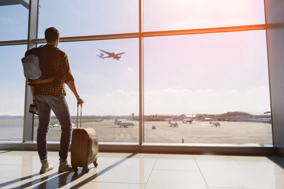 Calm male tourist is standing in airport and looking at aircraft flight through window. He is holding tickets and suitcase. Sunset