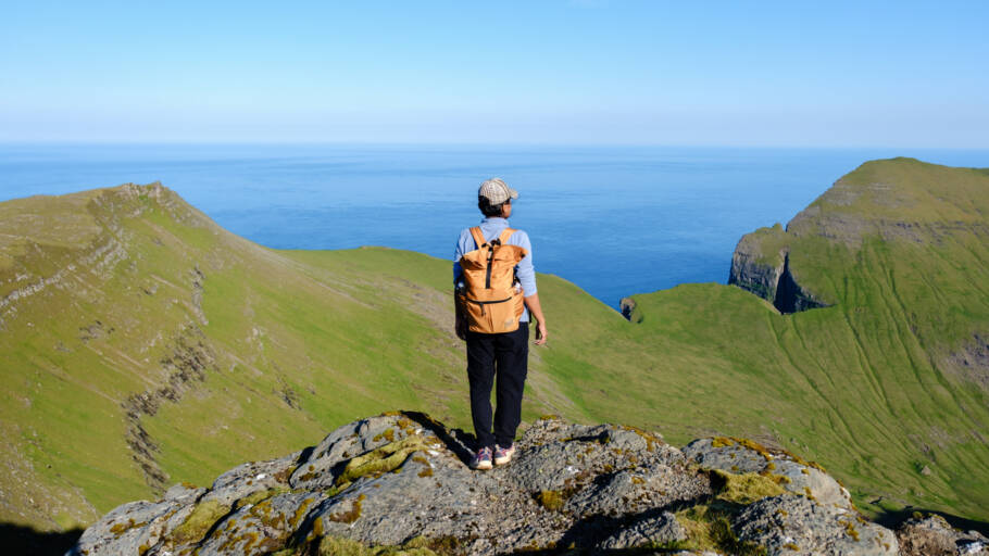 A solitary traveler gazes at the breathtaking coastal landscape of the Faroe Islands, surrounded by vibrant greenery and calm blue waters under a bright sky. The beauty is truly mesmerizing.