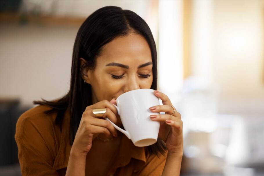 Mulher com cabelo liso preto curto, usando camiseta marrom e tomando chá em caneca branca usando anel dourado em um dos dedos
