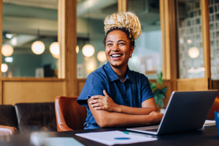 A cheerful young woman with blonde braids is seated at a desk in an office, smiling warmly. Her workspace includes a laptop, papers, and a pleasant background showcasing soft light and open spaces.