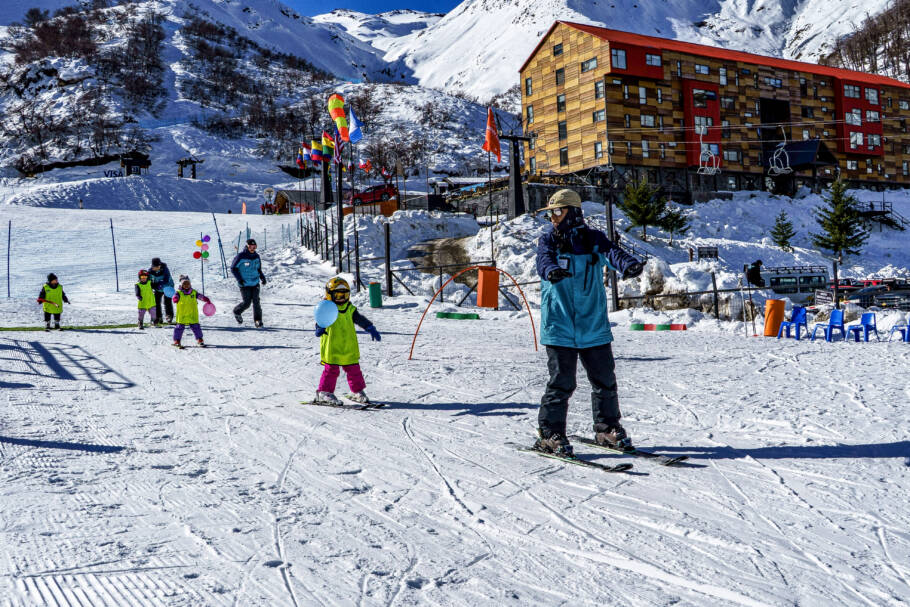 Crianças participam de aulas coletivas na base da montanha de Nevados de Chillán com hotel Alto Nevados ao fundo