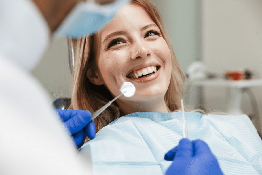 Uma jovem com cabelo loiro está sentada em uma cadeira odontológica, sorrindo enquanto recebe atendimento. Um profissional, parcialmente visível em primeiro plano, usa luvas azuis e examina a boca dela com um espelho odontológico. A paciente veste um avental descartável azul-claro, e o consultório aparece desfocado ao fundo.