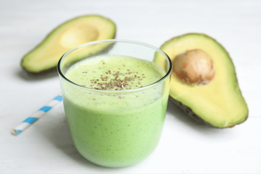 Copo transparente com vitamina verde coberta com sementes de chia, posicionado sobre uma superfície branca ao lado de metades de abacate e um canudo listrado de azul e branco.