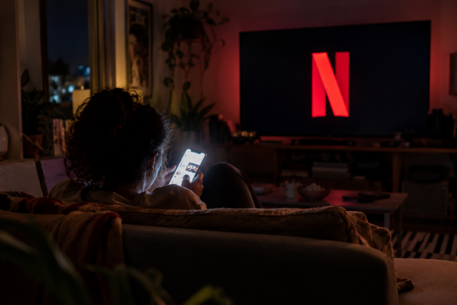 Espectadora divide a atenção entre a Netflix e o celular. A dispersão durante o consumo de filmes e séries pode afetar a formação de memórias e a criação de fãs (Imagem gerada por IA)